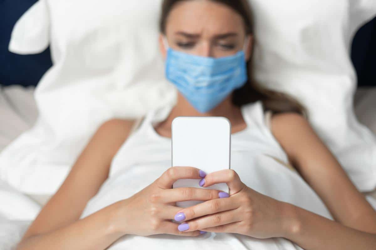 Girl In Mask Using Smartphone Lying In Bed At Home